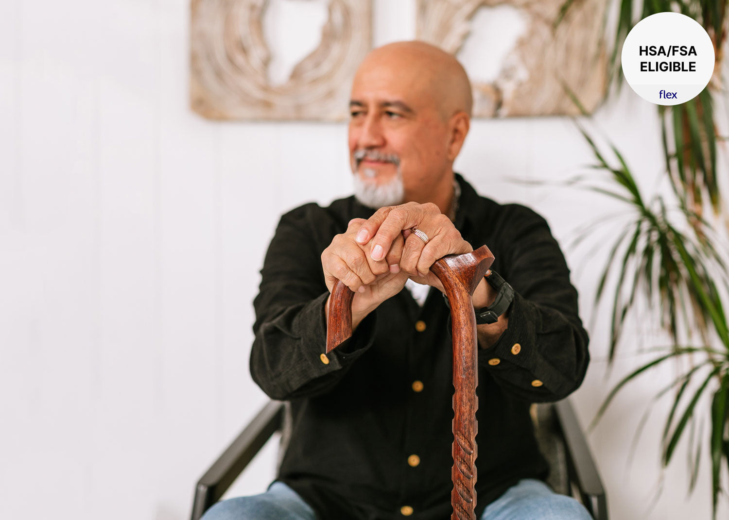 Man sitting in a chair holding a wooden cane with a plant and decorative wall piece in the background.